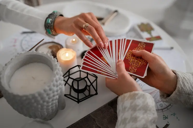Woman's hands fanning red tarot cards with gold design, lit candle and astrology chart in background - Roosy Spirit Melbourne